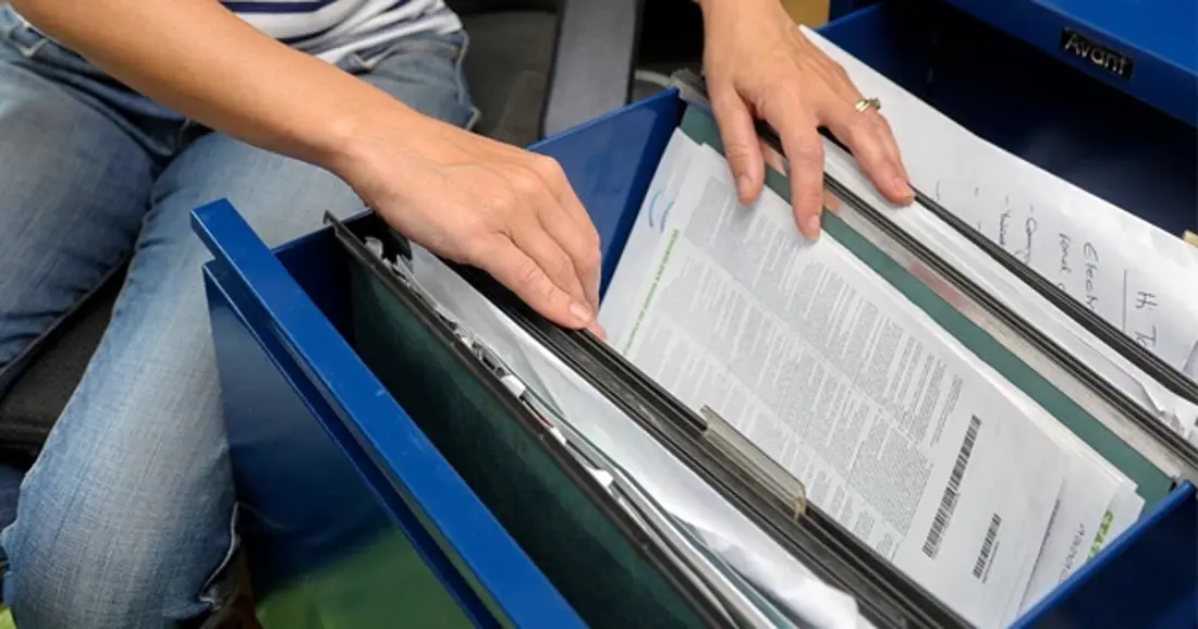 A person looking through an open filing cabinet drawer with suspended files and paperwork.