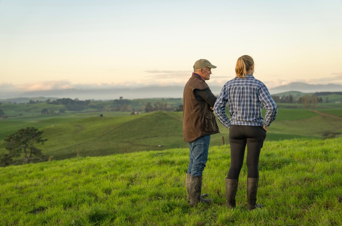 A male and a female dairy farmer standing in an open paddock overlooking the farm.