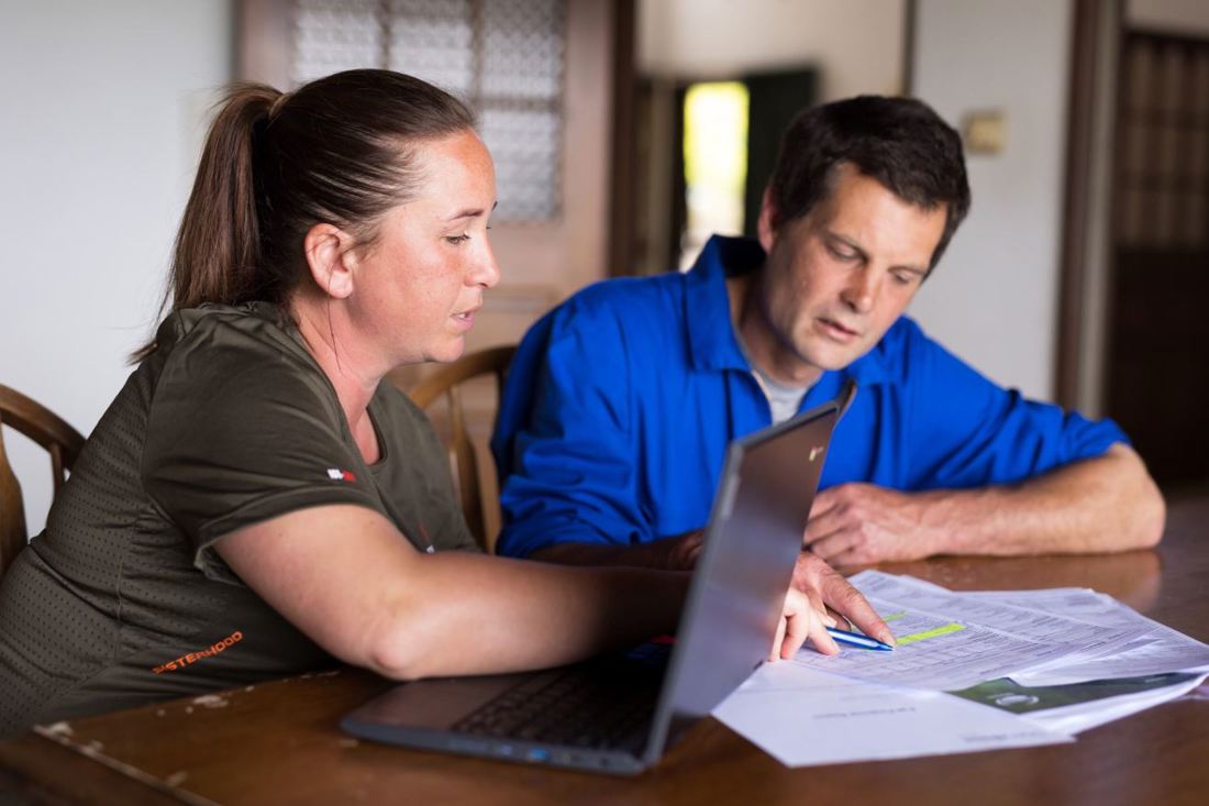 New Zealand dairy farmers Danielle and Bevan Cornelius sitting at a desk monitoring their farm expenses using a laptop and printed data.